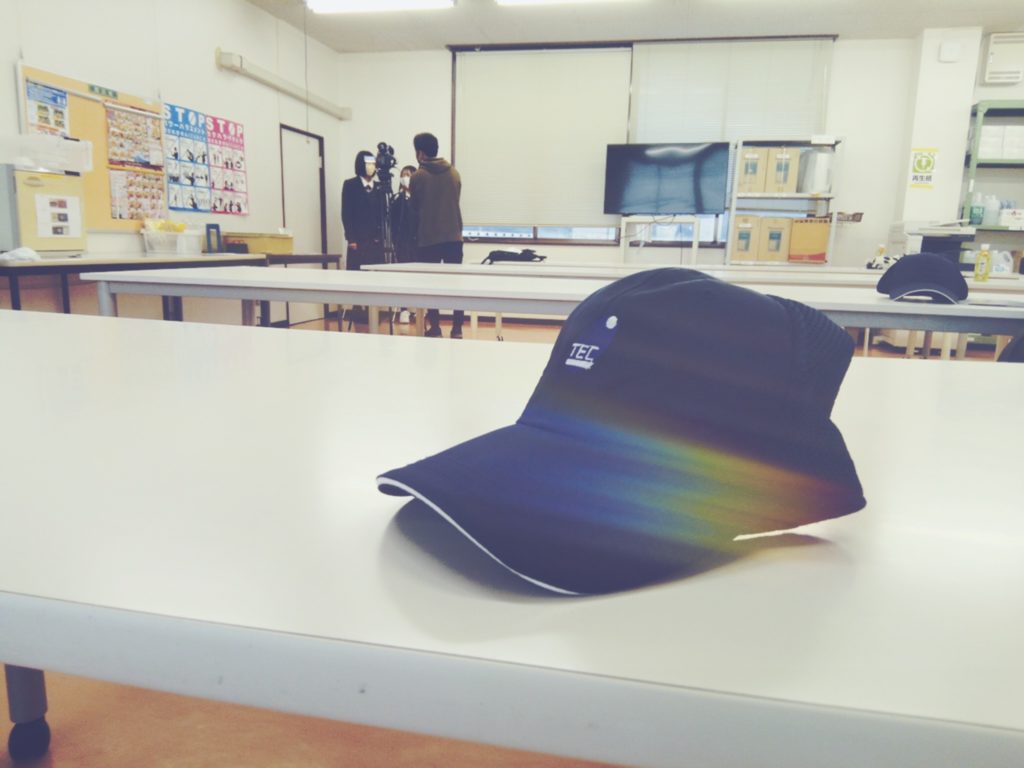 東洋電制製作所のロゴが入った紺色の帽子と、NET3のインタビューを受ける生徒2人の様子